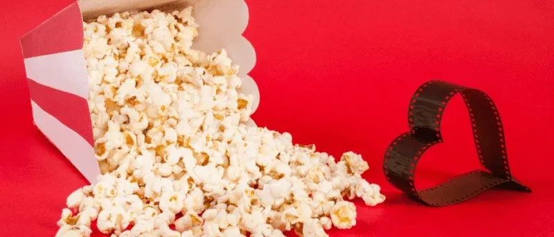 Spilled popcorn from a red and white box next to a heart-shaped film strip on a red background.